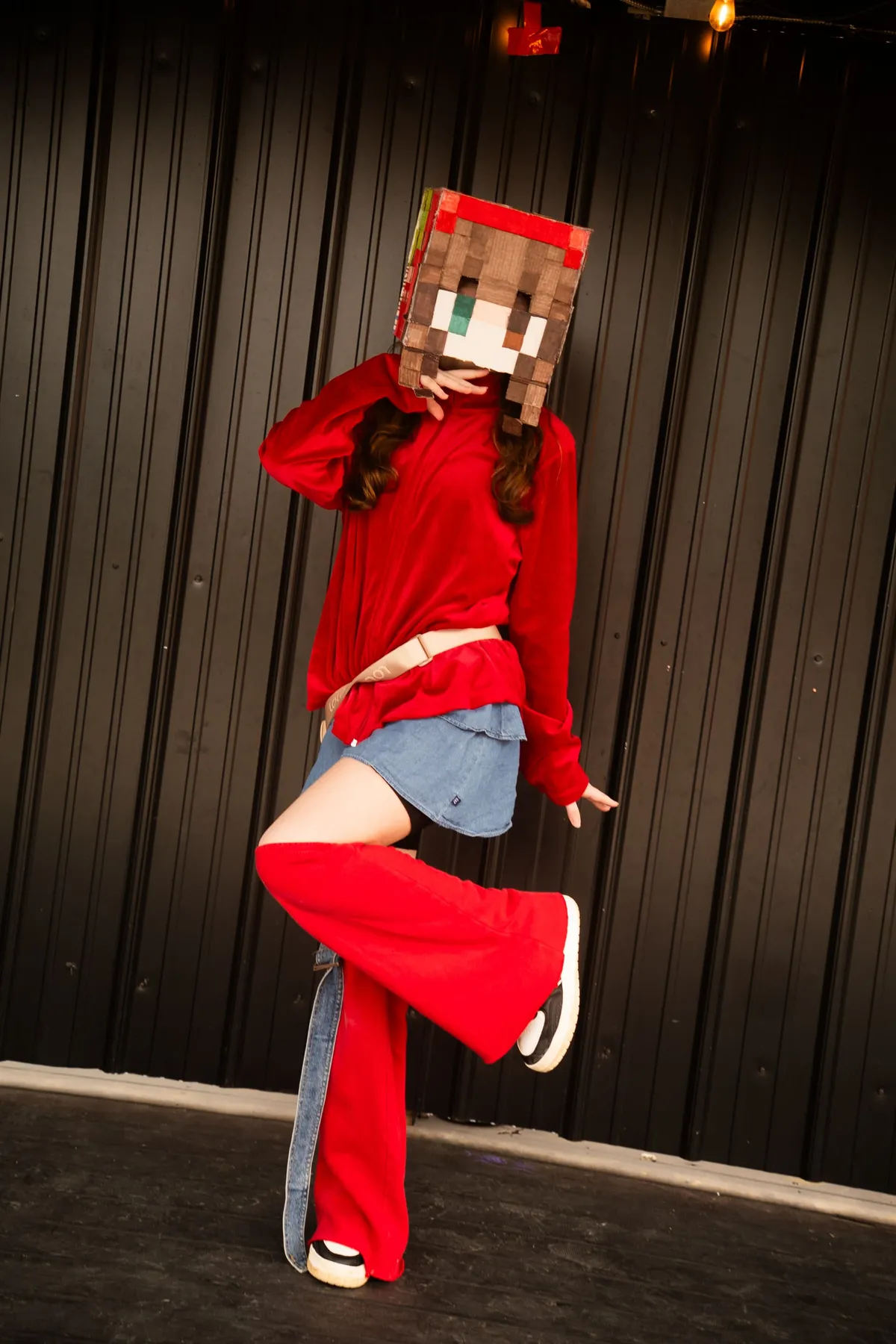 A cosplayer wearing a handmade pixelated cardboard block head, red top, denim skirt and red leg warmers strikes a dance pose on stage at Knox Pop Con.